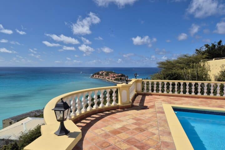 Terrace overlooking the blue sea with a yellow balustrade and terracotta tiles, a pool on the right, and a sunny sky above.