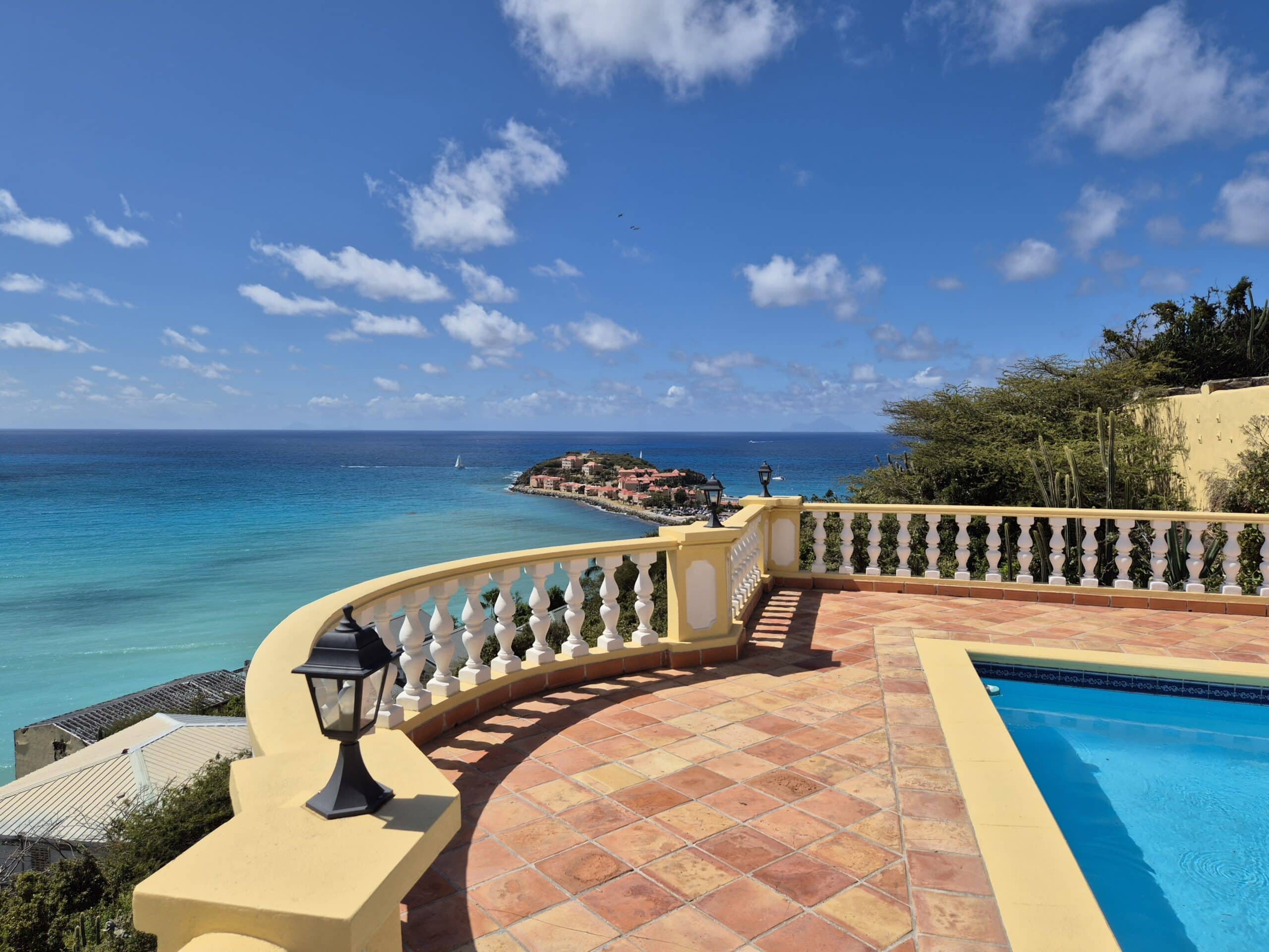 Terrace overlooking the blue sea with a yellow balustrade and terracotta tiles, a pool on the right, and a sunny sky above.