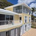 Yellow and gray coastal apartment building with white railings and open balconies under a blue sky, hillside in the background.