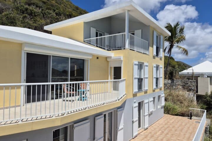 Yellow and gray coastal apartment building with white railings and open balconies under a blue sky, hillside in the background.