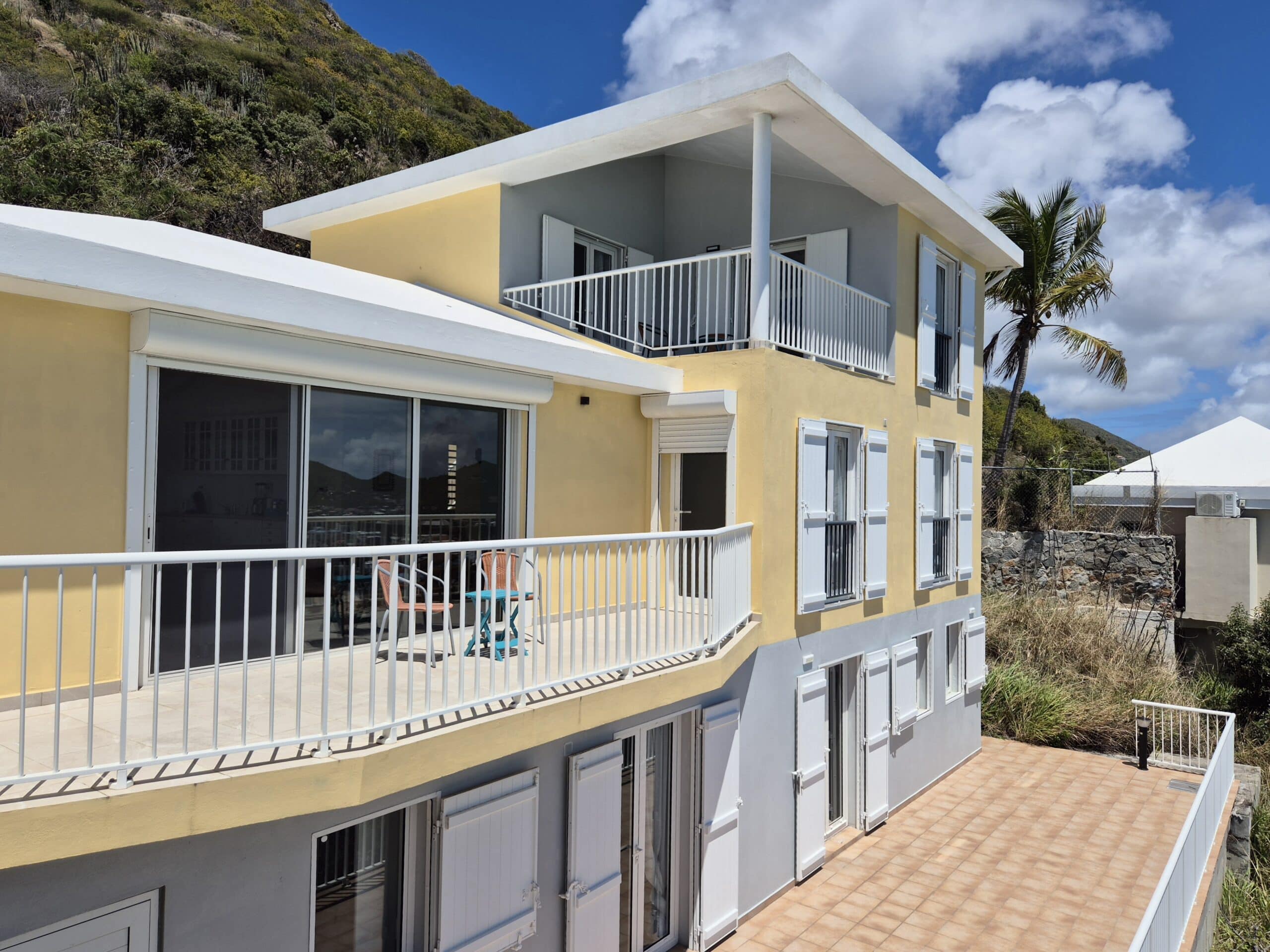 Yellow and gray coastal apartment building with white railings and open balconies under a blue sky, hillside in the background.