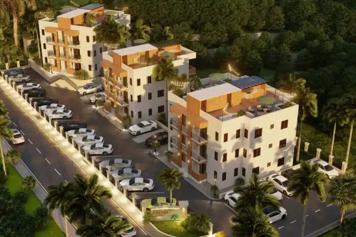 Aerial view of a modern, multi-building apartment complex with white and tan façades, palm trees, string lights, and a curved row of parked cars along a gated entrance.