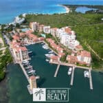 Aerial view of a coastal resort with multiple white and beige buildings along a marina, red-tiled roofs, and turquoise water.