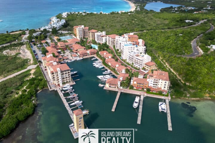 Aerial view of a coastal resort with multiple white and beige buildings along a marina, red-tiled roofs, and turquoise water.