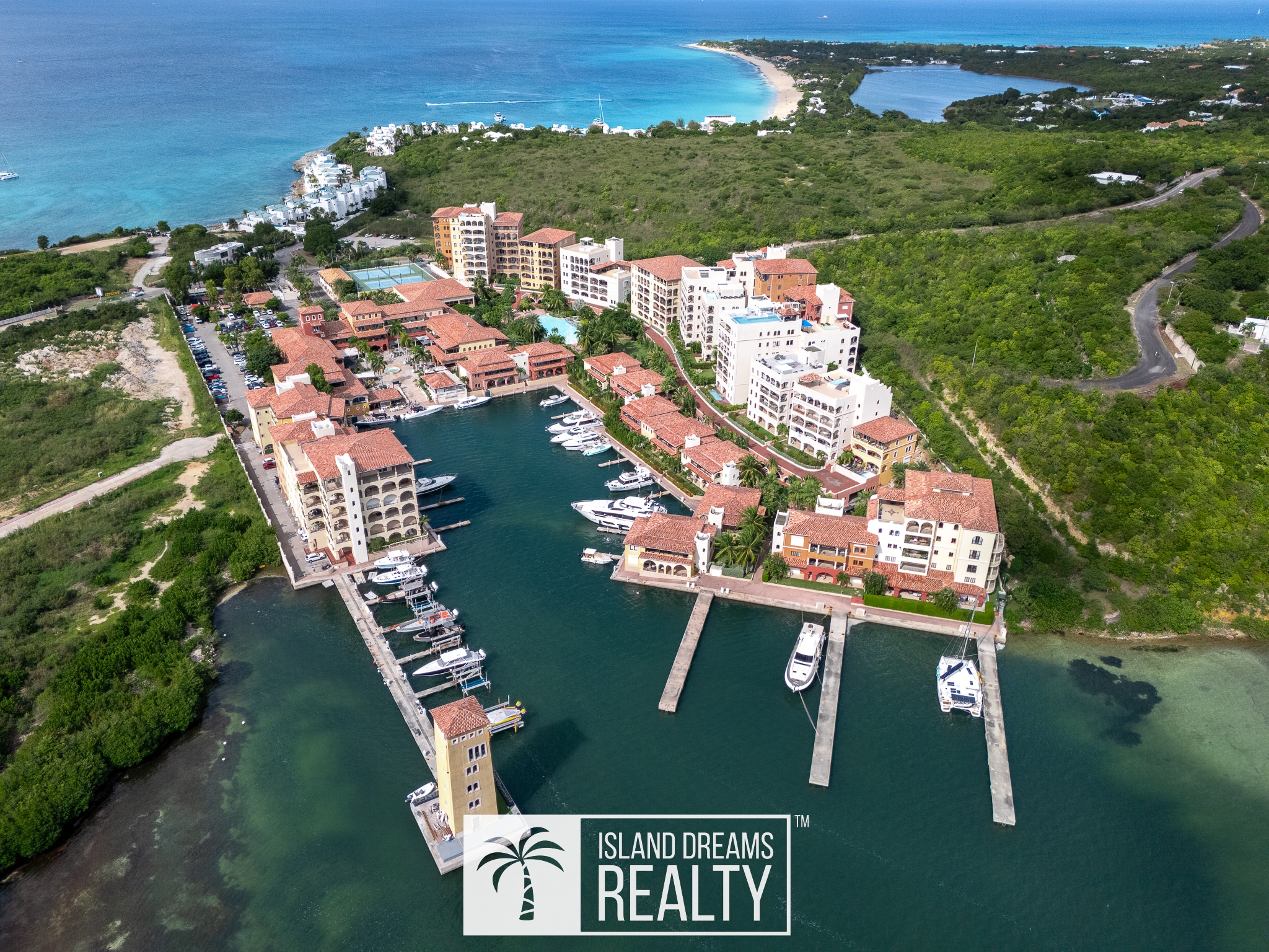 Aerial view of a coastal resort with multiple white and beige buildings along a marina, red-tiled roofs, and turquoise water.