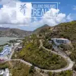 Aerial view of a hillside residential area with winding road, low buildings, and a body of water on the left under a blue sky with clouds. Waterfront community surrounded by dry hills.