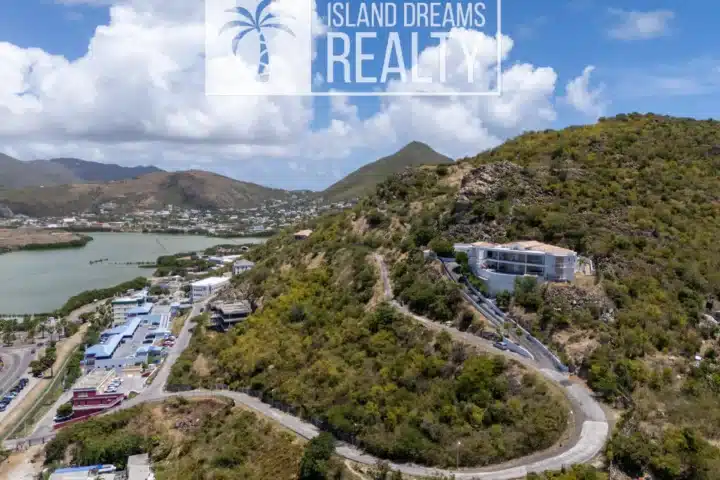 Aerial view of a hillside residential area with winding road, low buildings, and a body of water on the left under a blue sky with clouds. Waterfront community surrounded by dry hills.