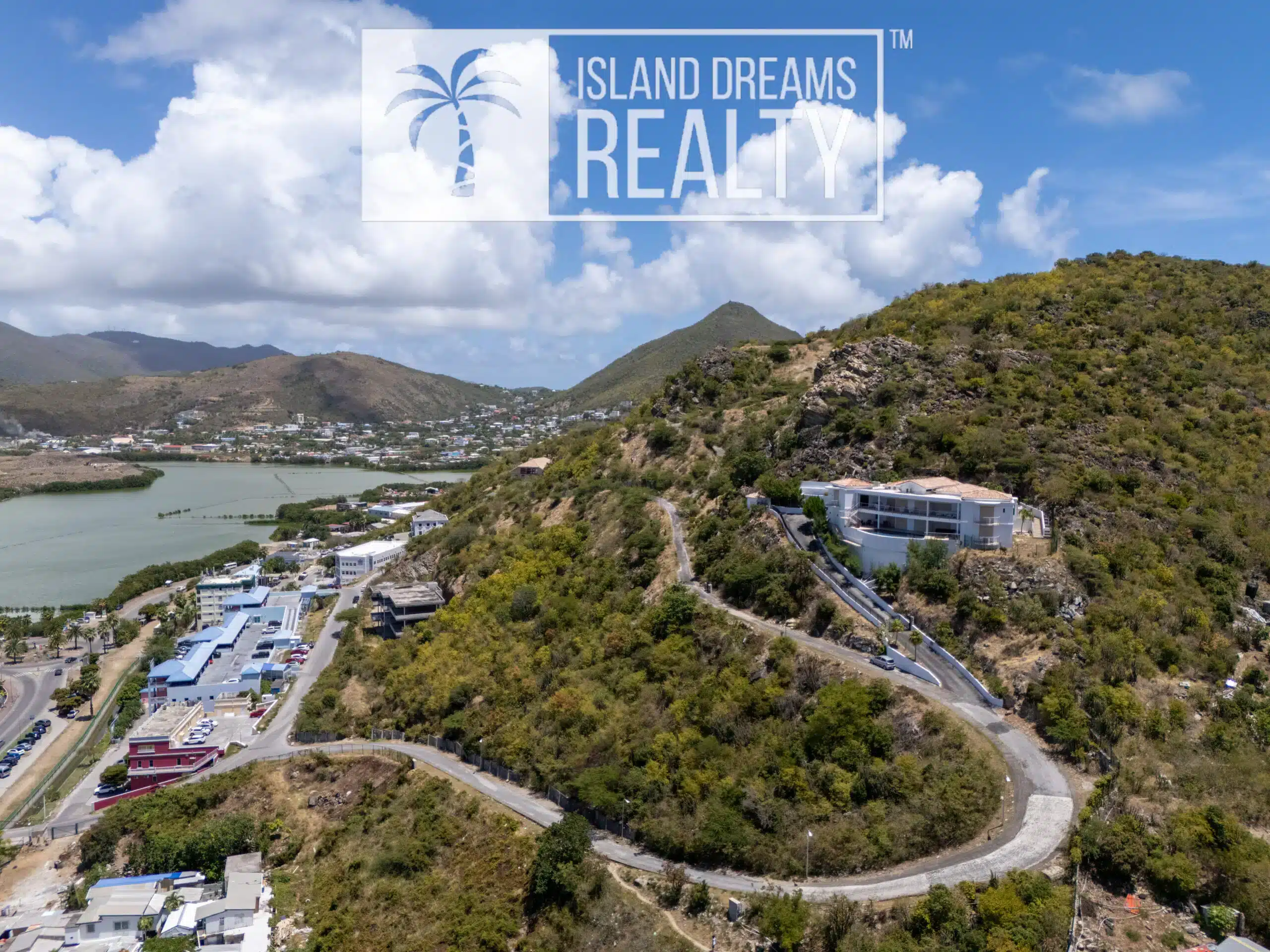 Aerial view of a hillside residential area with winding road, low buildings, and a body of water on the left under a blue sky with clouds. Waterfront community surrounded by dry hills.