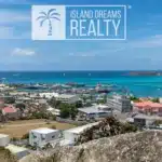 Coastal cityscape with turquoise bay, boats, and colorful buildings; a large 'Island Dreams Realty' logo overlays the sky above the scene.