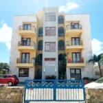 Four-story apartment building with curved balconies, a blue central column, palm trees, and a blue gate at the entrance, 'Ocean's' sign visible.