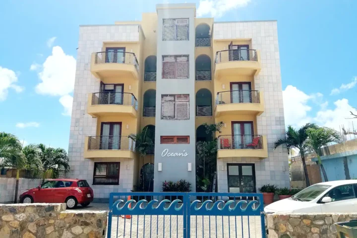 Four-story apartment building with curved balconies, a blue central column, palm trees, and a blue gate at the entrance, 'Ocean's' sign visible.