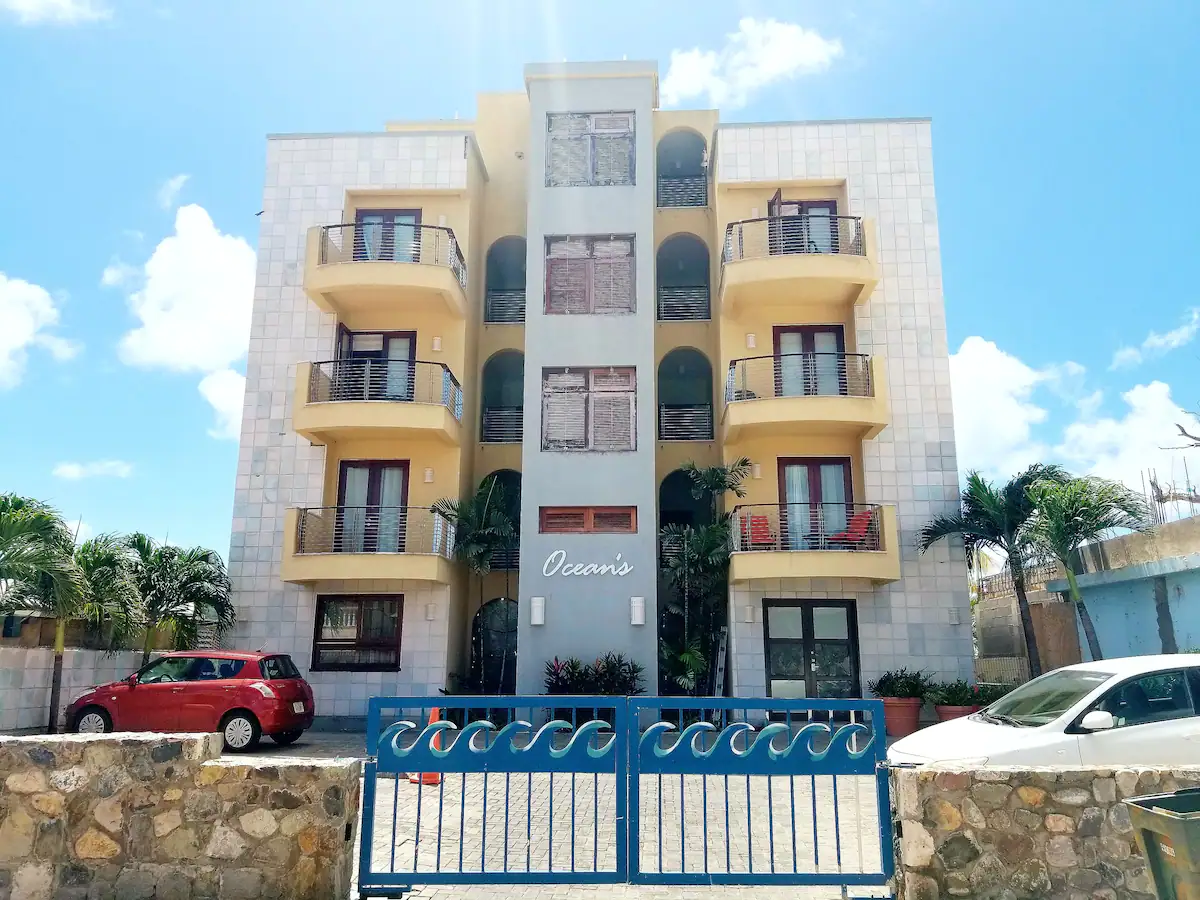 Four-story apartment building with curved balconies, a blue central column, palm trees, and a blue gate at the entrance, 'Ocean's' sign visible.