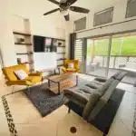 Modern living room with gray sectional sofa, two mustard-yellow lounge chairs, and a wooden coffee table on a gray rug. A wall unit with shelves and a mounted TV are on one wall, and large sliding doors open to a patio with outdoor seating.