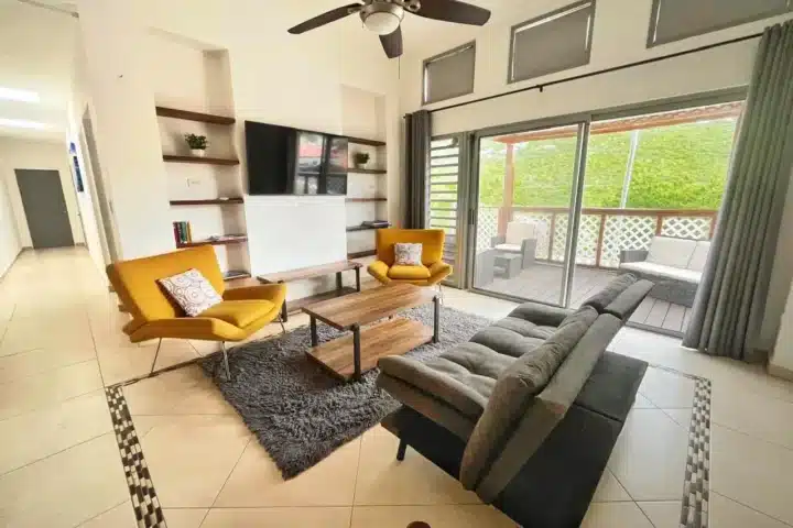 Modern living room with gray sectional sofa, two mustard-yellow lounge chairs, and a wooden coffee table on a gray rug. A wall unit with shelves and a mounted TV are on one wall, and large sliding doors open to a patio with outdoor seating.