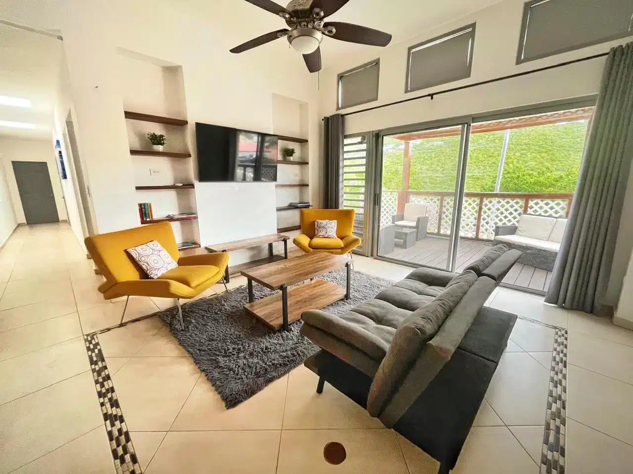 Modern living room with gray sectional sofa, two mustard-yellow lounge chairs, and a wooden coffee table on a gray rug. A wall unit with shelves and a mounted TV are on one wall, and large sliding doors open to a patio with outdoor seating.