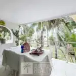 Covered outdoor patio with a white tablecloth, potted plant, and a decorative birdcage on a table; tropical plants and trees visible beyond.