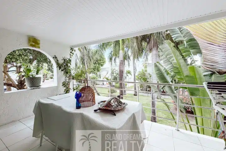 Covered outdoor patio with a white tablecloth, potted plant, and a decorative birdcage on a table; tropical plants and trees visible beyond.