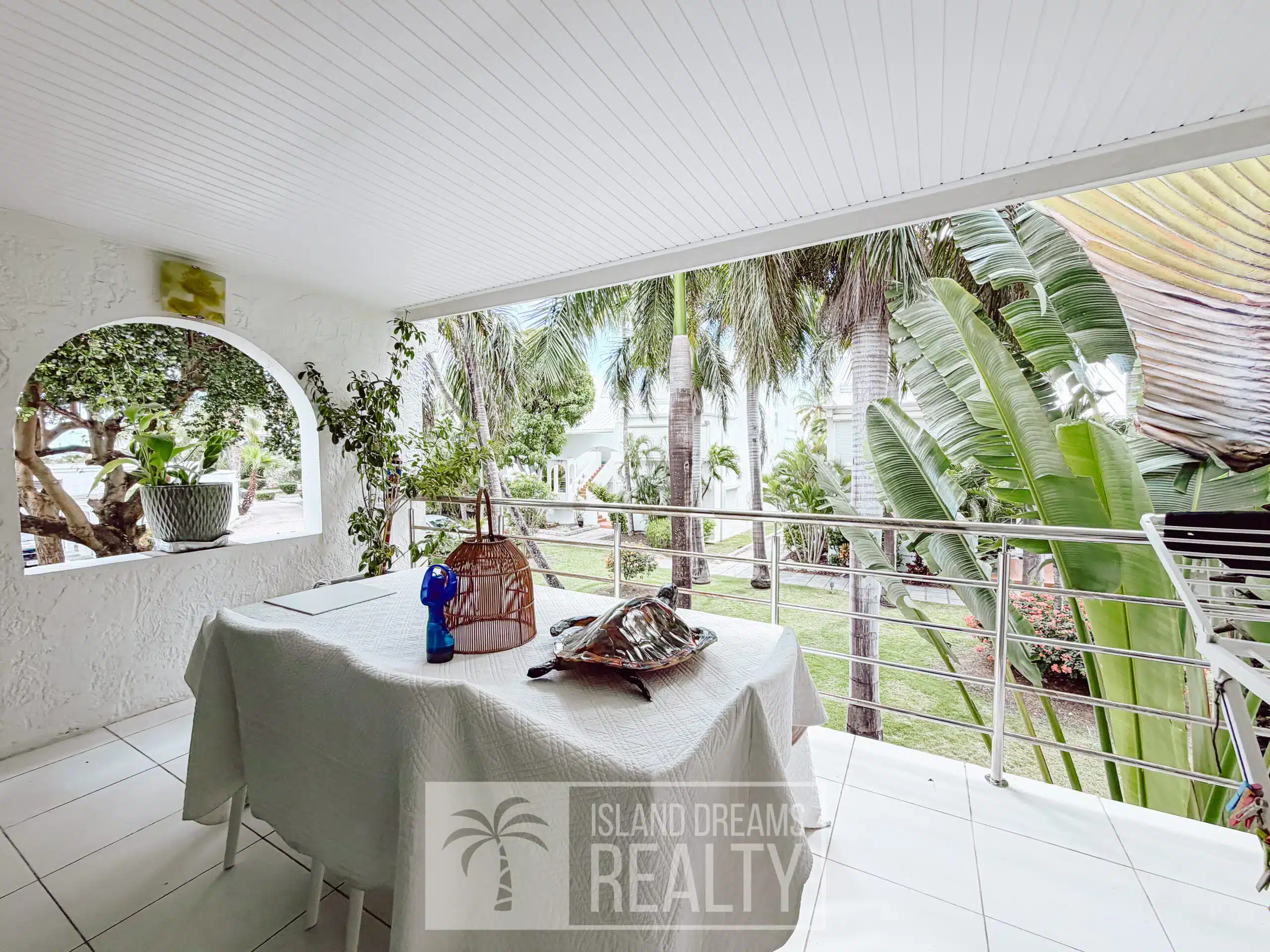 Covered outdoor patio with a white tablecloth, potted plant, and a decorative birdcage on a table; tropical plants and trees visible beyond.