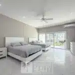 Bright modern bedroom with two white platform beds, gray bedding, white nightstands, and a ceiling fan; sliding glass doors to a sunny balcony.