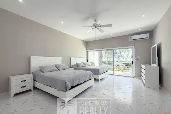 Bright modern bedroom with two white platform beds, gray bedding, white nightstands, and a ceiling fan; sliding glass doors to a sunny balcony.