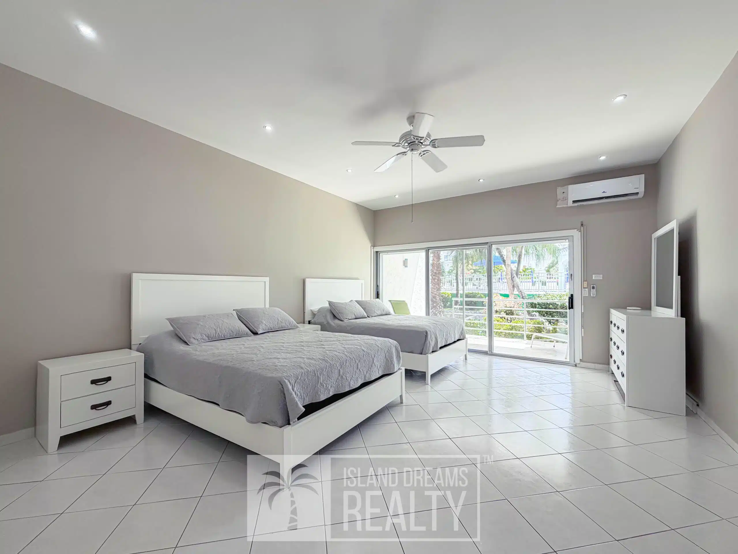 Bright modern bedroom with two white platform beds, gray bedding, white nightstands, and a ceiling fan; sliding glass doors to a sunny balcony.