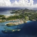 Aerial view of a tropical island with a curved shoreline, turquoise bays, and a town of white and red-roofed buildings along the coast.