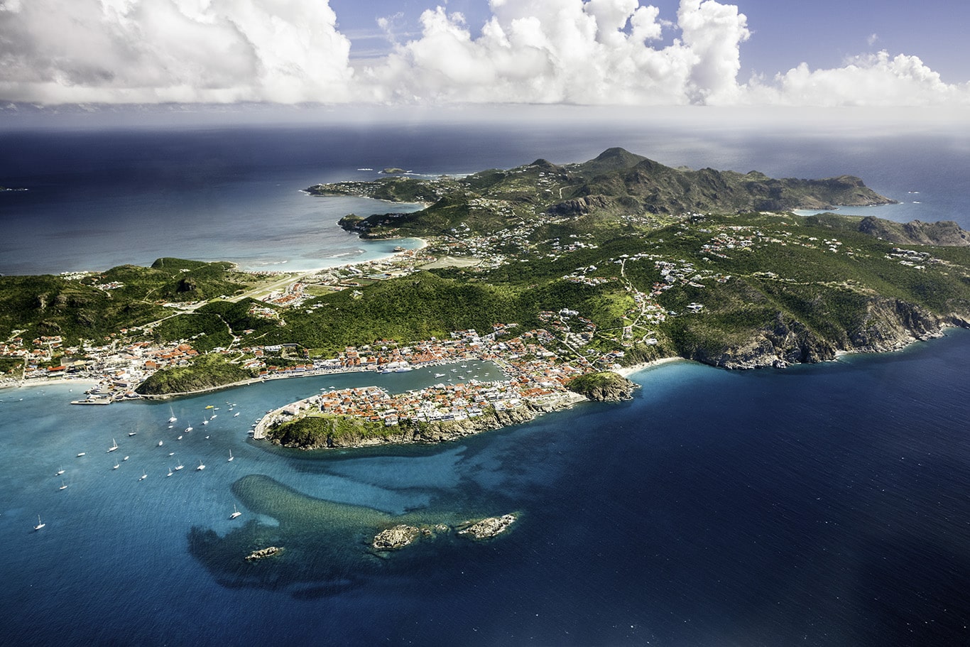 Aerial view of a tropical island with a curved shoreline, turquoise bays, and a town of white and red-roofed buildings along the coast.