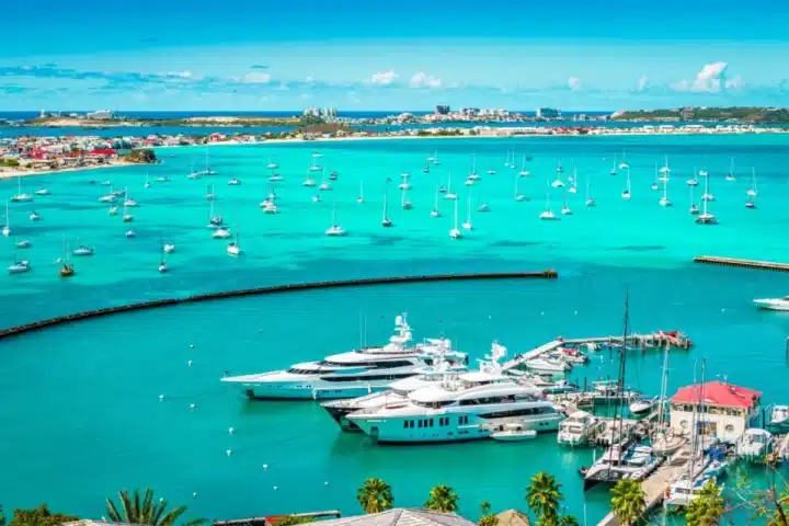 Turquoise harbor filled with moored sailboats and luxury yachts at a sunny marina, with palm trees in the foreground.