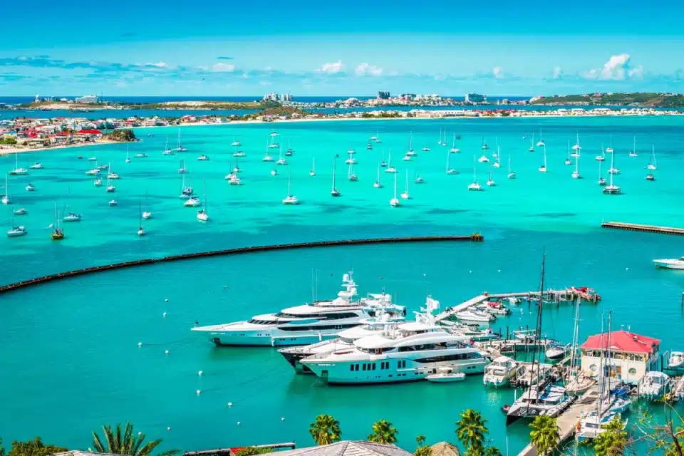 Turquoise harbor filled with moored sailboats and luxury yachts at a sunny marina, with palm trees in the foreground.