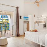 Bright hotel suite with a white bed, breakfast tray, and a kitchenette, open to a balcony with blue railing and outdoor seating.