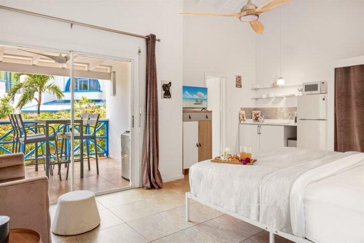 Bright hotel suite with a white bed, breakfast tray, and a kitchenette, open to a balcony with blue railing and outdoor seating.