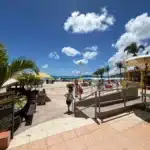 Sunny beach scene with palm trees, colorful umbrellas, and a yellow bar near the shore.