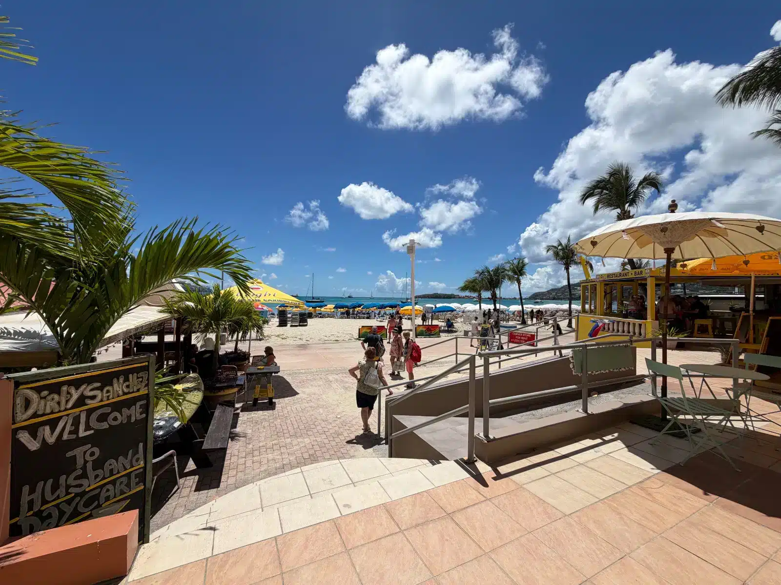 Sunny beach scene with palm trees, colorful umbrellas, and a yellow bar near the shore.
