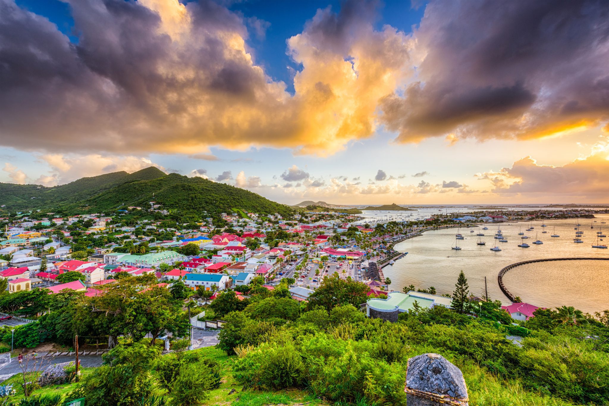 Colorful hillside town by the sea with boats in a sheltered harbor at sunsetfall, cloud-filled sky above.