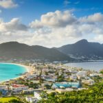 Coastal town with turquoise bay, colorful buildings, and surrounding mountains under a partly cloudy sky.