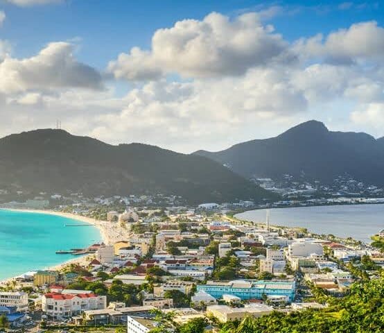 Coastal town with turquoise bay, colorful buildings, and surrounding mountains under a partly cloudy sky.