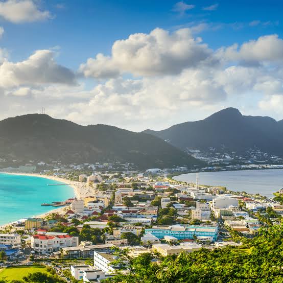 Coastal town with turquoise bay, colorful buildings, and surrounding mountains under a partly cloudy sky.