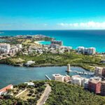 Aerial view of a coastal town with turquoise sea, white apartment blocks, and green vegetation along a peninsula.