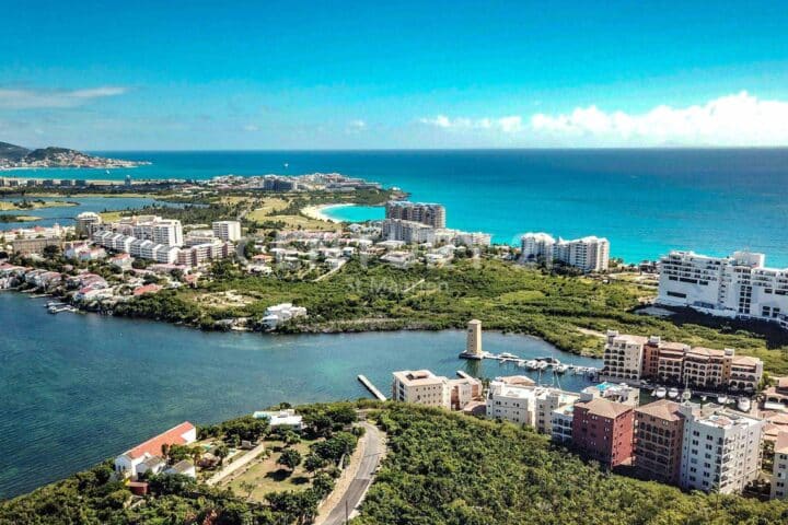 Aerial view of a coastal town with turquoise sea, white apartment blocks, and green vegetation along a peninsula.