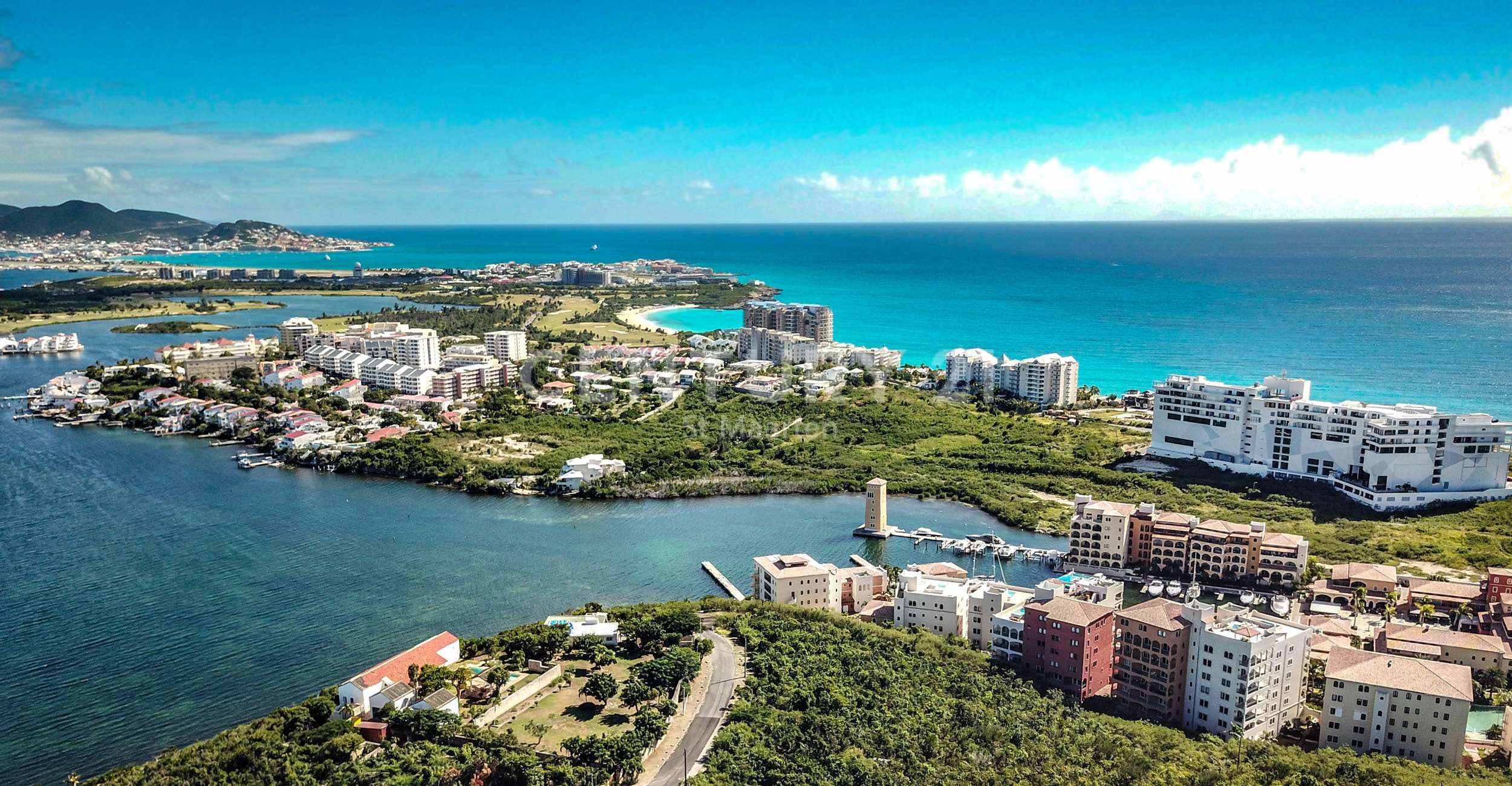 Aerial view of a coastal town with turquoise sea, white apartment blocks, and green vegetation along a peninsula.
