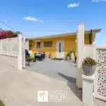 Yellow bungalow with white decorative gate and courtyard, outdoor seating, and potted plants under a clear blue sky (real estate listing).