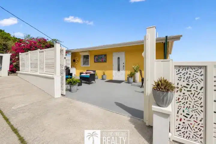 Yellow bungalow with white decorative gate and courtyard, outdoor seating, and potted plants under a clear blue sky (real estate listing).
