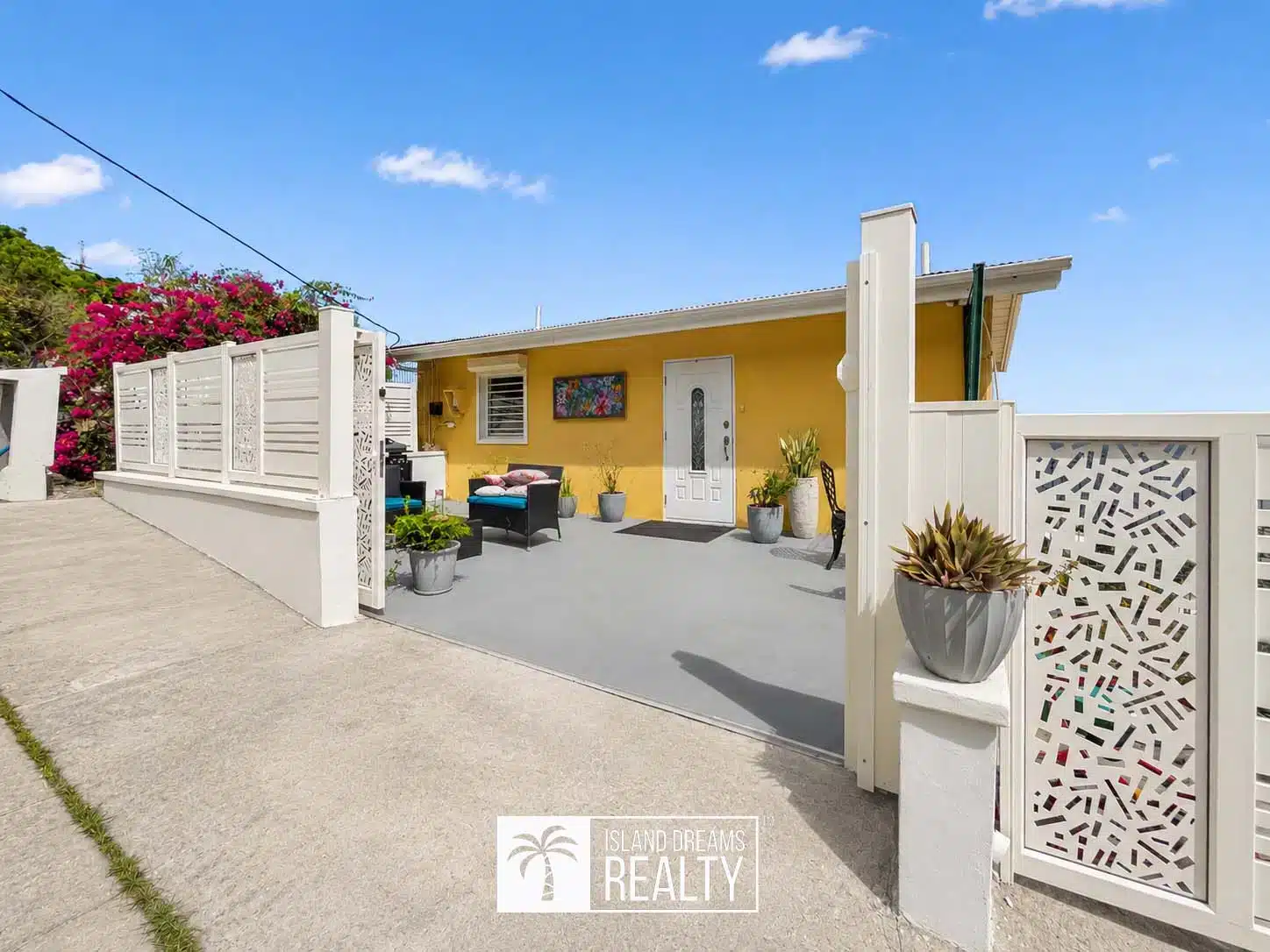 Yellow bungalow with white decorative gate and courtyard, outdoor seating, and potted plants under a clear blue sky (real estate listing).