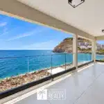 Balcony with glass railing overlooking a rocky coastline and blue sea under a bright sky.