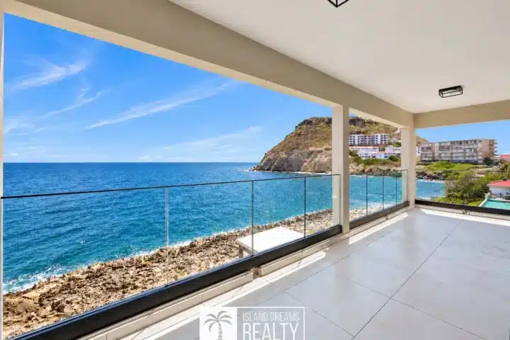 Balcony with glass railing overlooking a rocky coastline and blue sea under a bright sky.