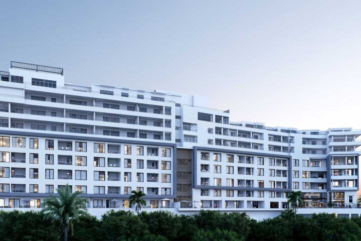 Modern white apartment building with many balconies and large windows, set against a clear blue sky with greenery in the foreground