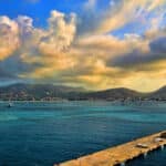 Pier extends into calm blue sea with a coastal town and mountains on the horizon under a colorful, cloudy sky at sunset.