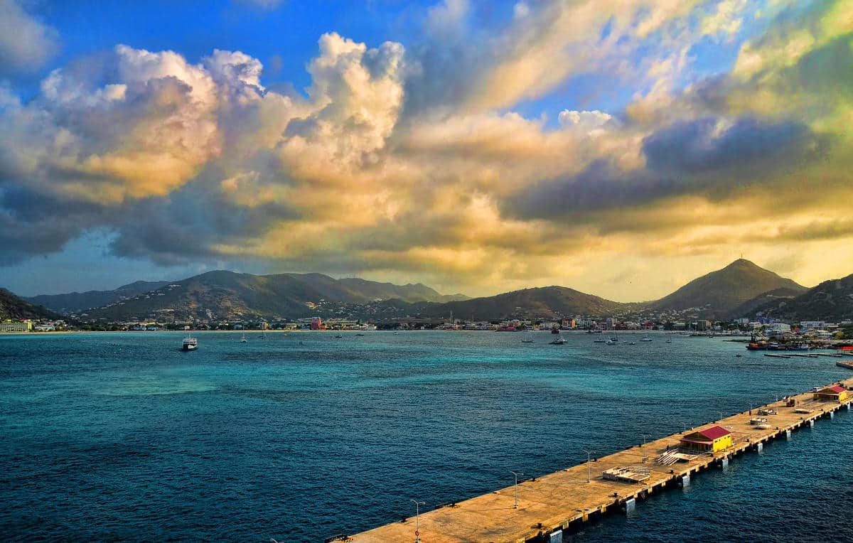 Pier extends into calm blue sea with a coastal town and mountains on the horizon under a colorful, cloudy sky at sunset.