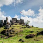 Ruined stone fortress on a grassy hill with a small flag atop, visitors near the ruins, and a woman in a hat walking with a pink bag under a blue sky.