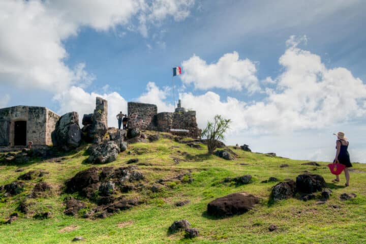 Ruined stone fortress on a grassy hill with a small flag atop, visitors near the ruins, and a woman in a hat walking with a pink bag under a blue sky.